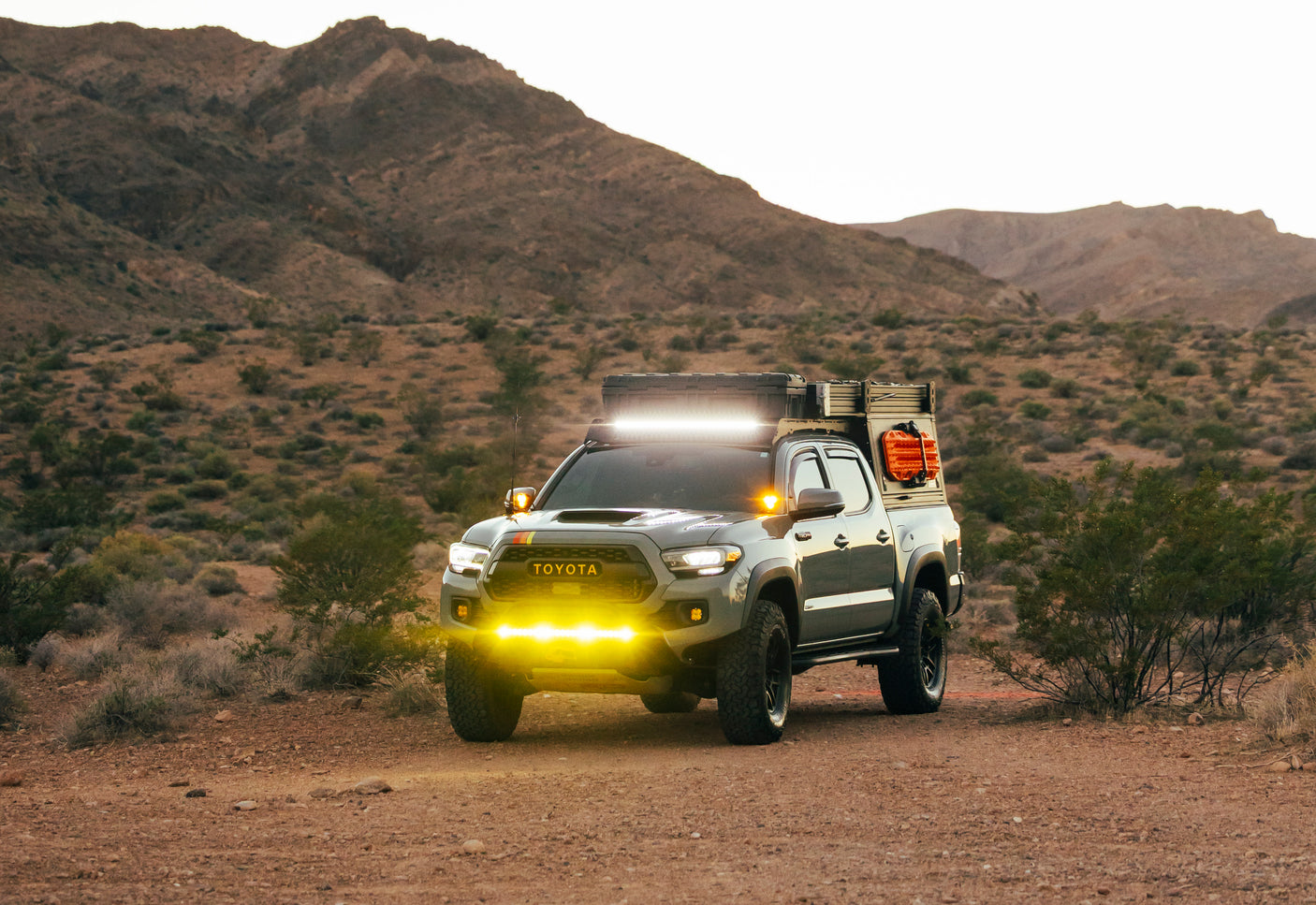 Rugged Toyota Tacoma overlanding truck with bright lights in desert mountains. Explore with this unique RV rental!