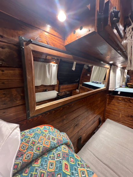 Inside an RV rental, a mirror reflects the cozy, wood-paneled interior featuring beds and a window with white curtains.