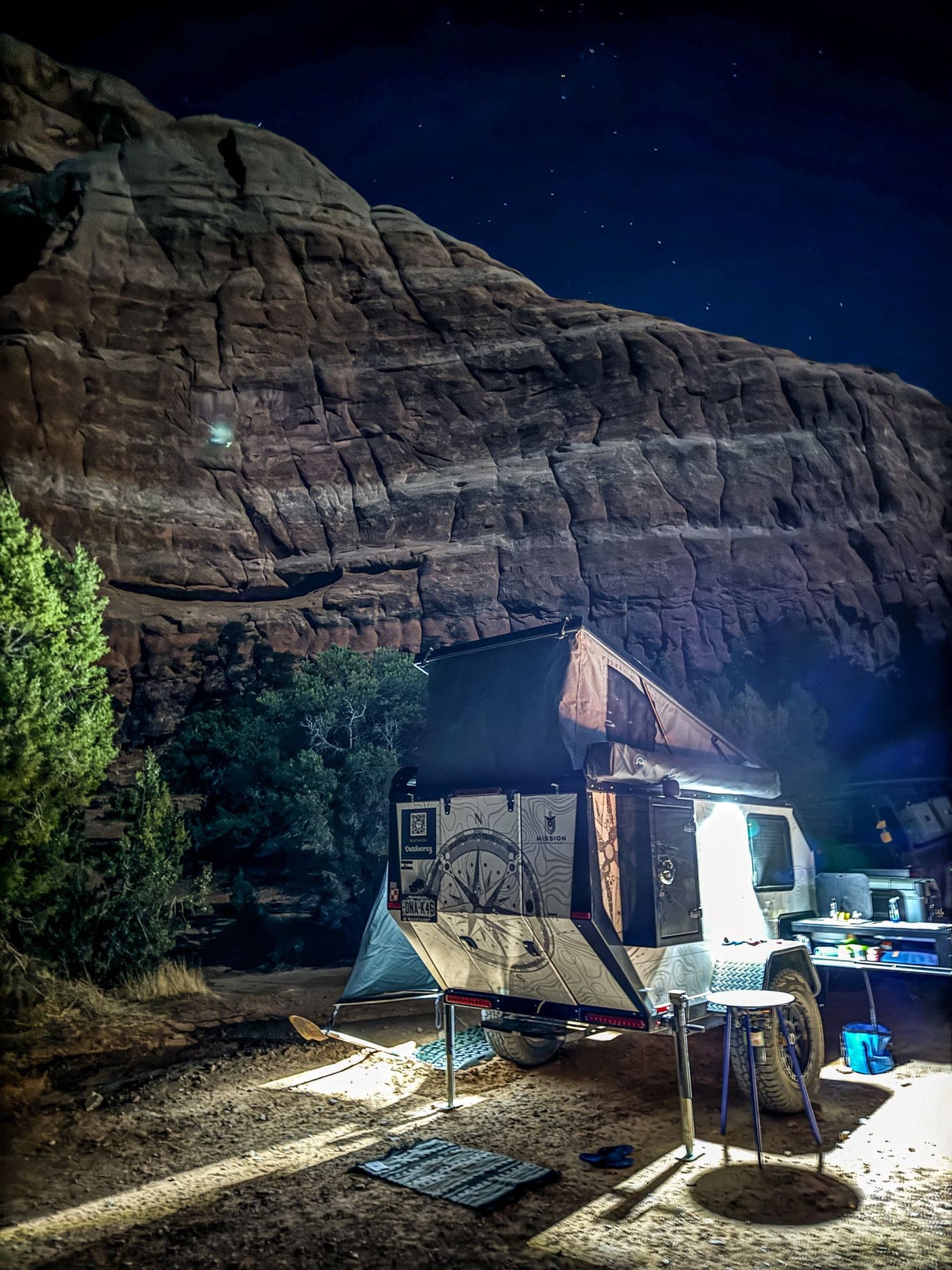 An RV rental trailer with a rooftop tent illuminated at night, parked before a massive rock face and starry sky.