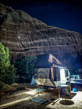 An RV rental trailer with a rooftop tent illuminated at night, parked before a massive rock face and starry sky.