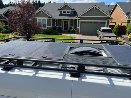 RV rental van's roof: black rack, solar panels, & MaxxAir fan. Ideal for mobile living, parked by a house.