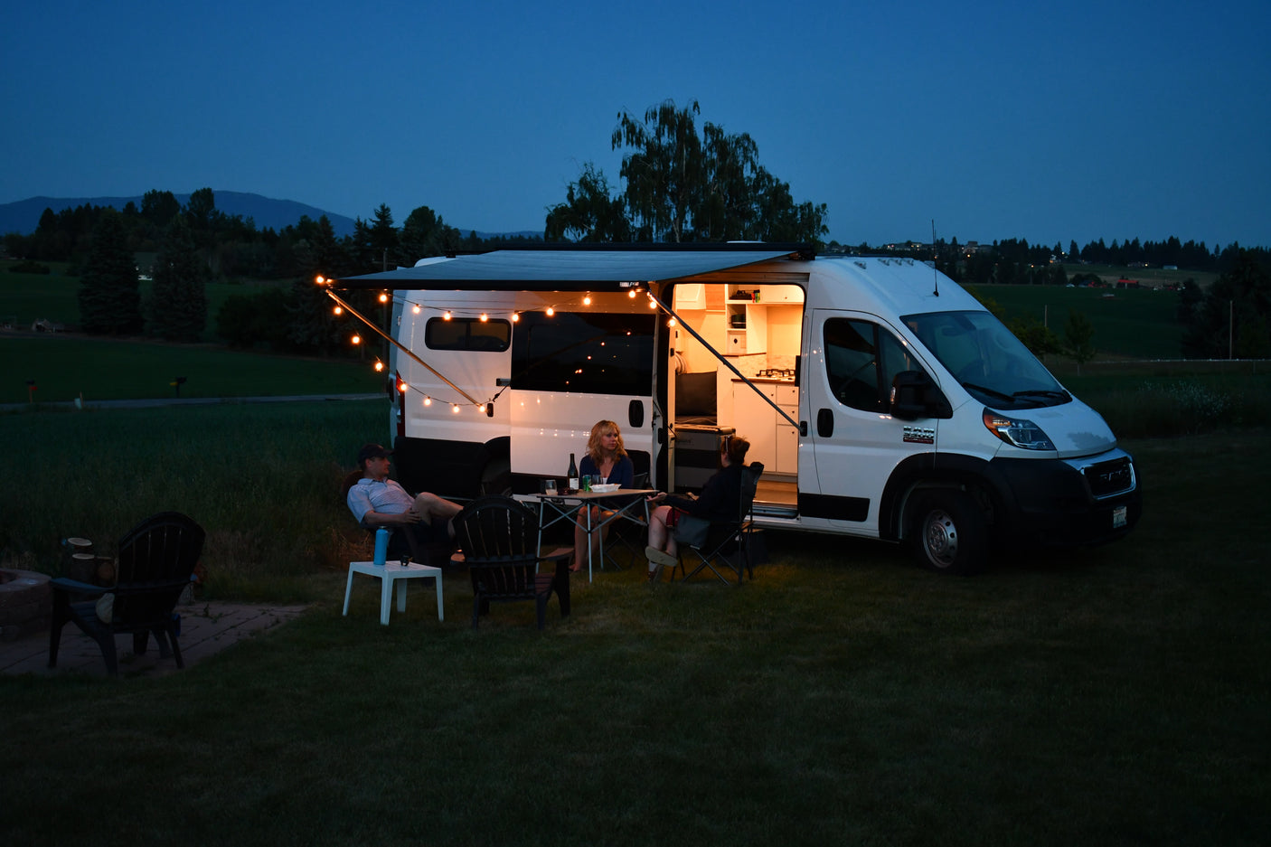 Perfect evening camping setup: Friends socializing outside a cozy RV rental camper van with string lights.