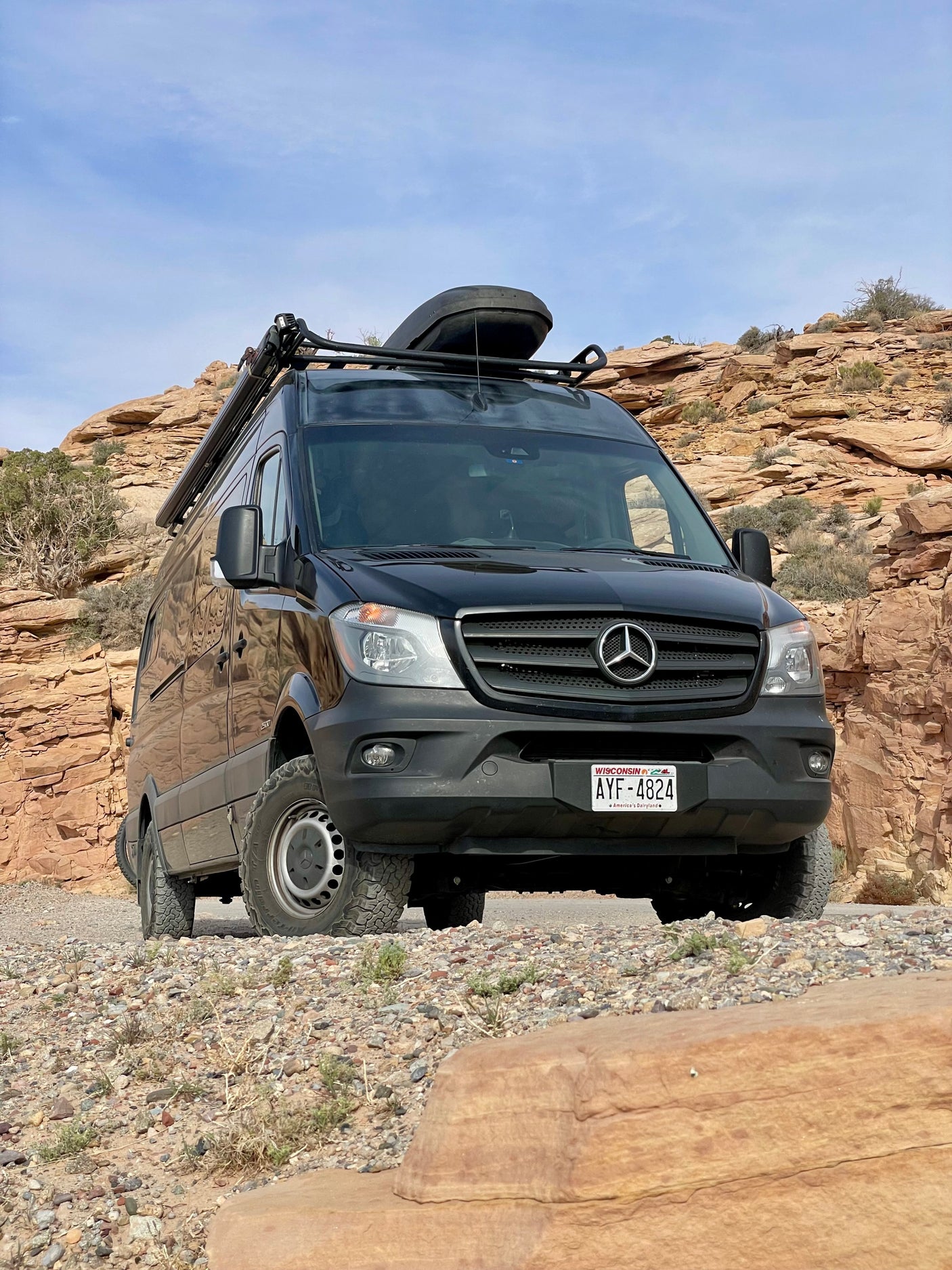 A black Mercedes-Benz Sprinter van equipped as an RV rental, ready for off-road adventures in a rocky desert landscape.