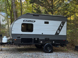 Grey and white Sun Lite LTD travel trailer, an ideal RV rental, parked on a gravel lot surrounded by lush trees.