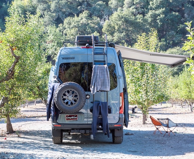 Grey Winnebago Revel RV rental parked in an orchard, ready for adventure. Awning extended, clothes drying on ladder, chairs set up. — photo 2
