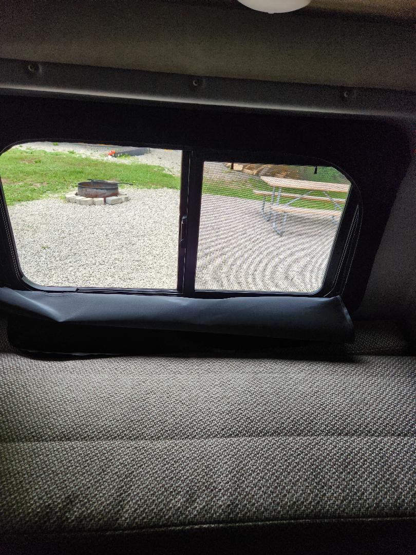 RV rental interior view: A window shows a campsite with a fire pit and picnic table, above comfortable gray bed cushions.