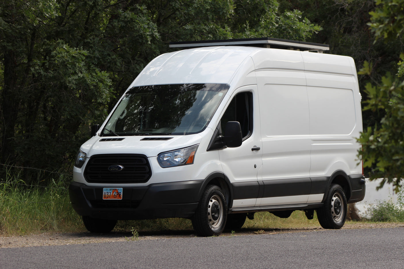 This white Ford Transit van with roof solar panels is ready for an RV rental adventure, parked on a road next to green trees.