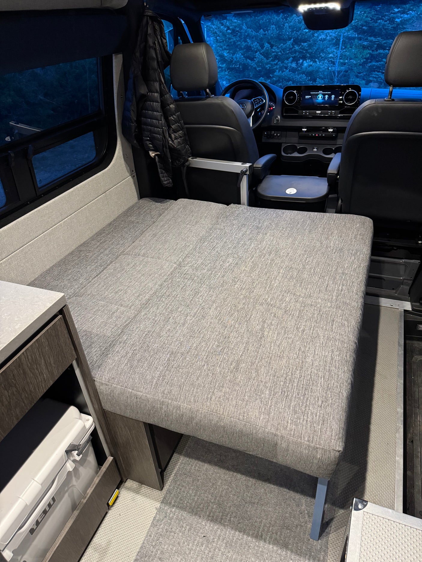 Interior of an rv rental camper van. A grey bed occupies the rear, with the driver's cockpit and a cooler visible.