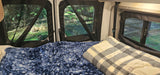 RV rental interior: cozy bed with blue blanket & plaid pillow, offering serene nature views through screened windows for camping.