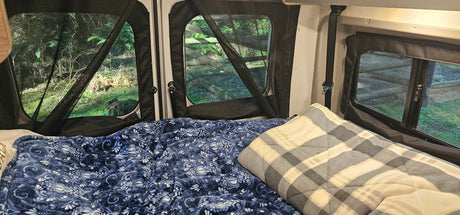 RV rental interior: cozy bed with blue blanket & plaid pillow, offering serene nature views through screened windows for camping.