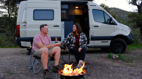 A happy couple holds hands by a warm campfire next to their Winnebago RV rental for a cozy camping trip.