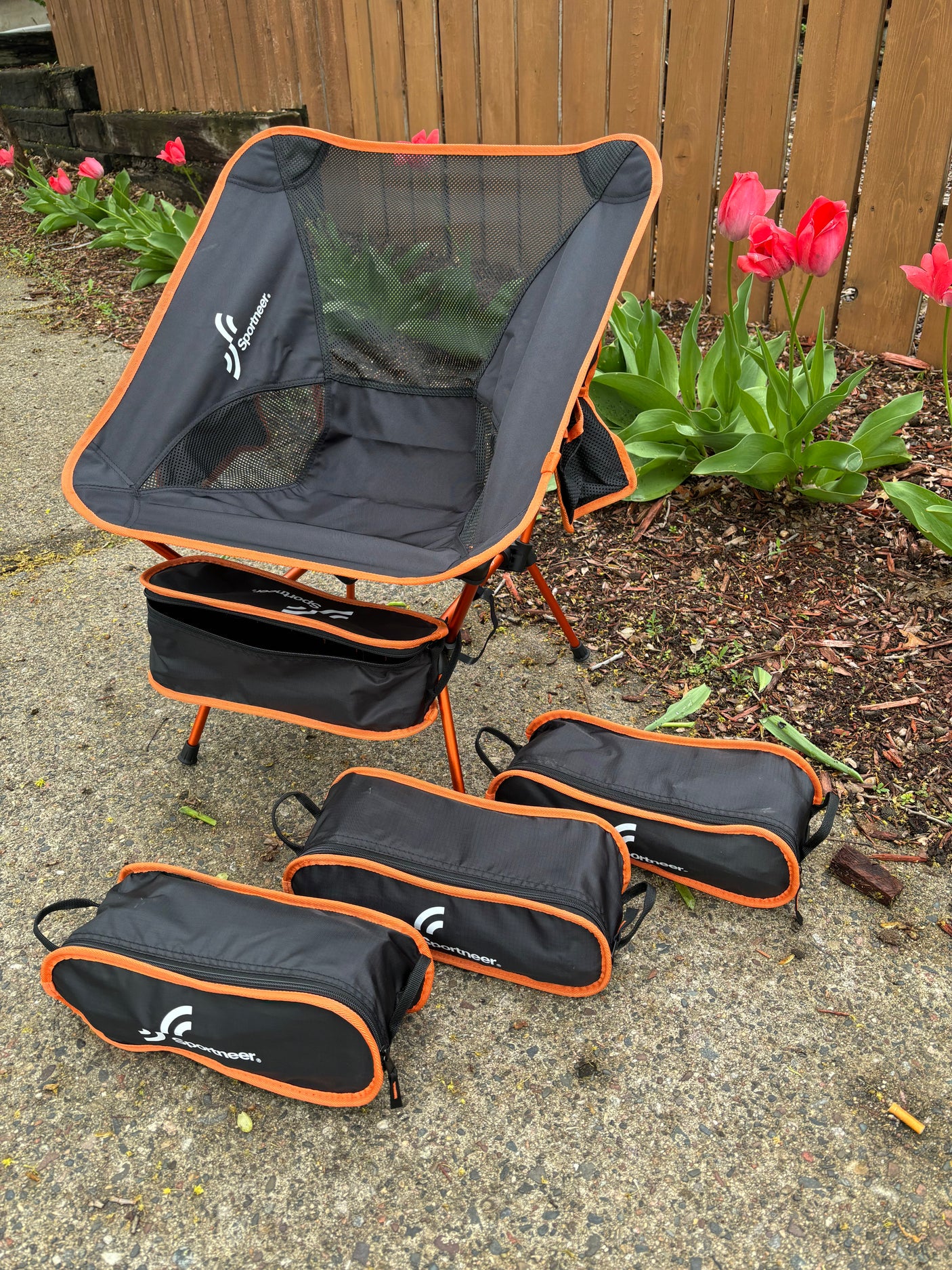 Sportneer portable chair & compact cases on pavement. Ideal outdoor gear for an RV rental trip, near tulips by a fence.
