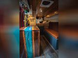 Inside a stylish custom RV rental: wood & blue resin kitchen, comfy bench seat, and bed. Perfect mobile living.
