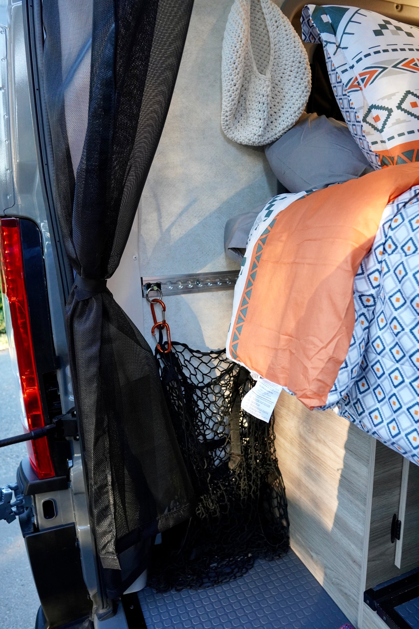 Cozy RV rental: patterned bedding, tied-back mesh screen, and storage net hanging from a wall track in a camper van.