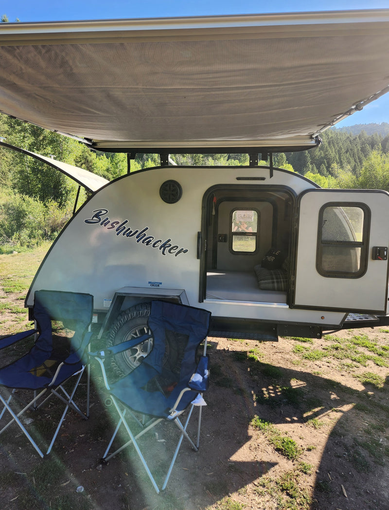 A white Bushwhacker teardrop RV rental with an extended awning and chairs set up for camping in a sunny mountain forest. — photo 8