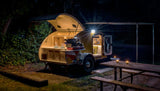 Cozy night camping in a small teardrop RV rental, with an open kitchen and lit interior, beside a picnic table.