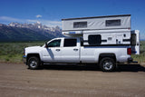 White pickup with Four Wheel pop-up camper, an ideal RV rental, parked on a dirt road by snow-capped mountains.