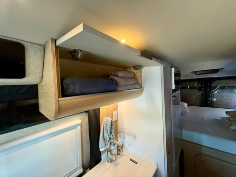 This RV rental features a bright, compact interior with open wood cabinets, neatly folded towels, a sink, and a comfortable bed.