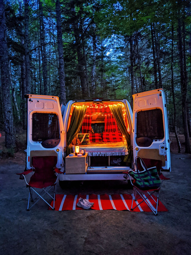 Cozy RV rental adventure: A camper van with glowing lights and open doors in a dark forest. Chairs & a rug sit outside.