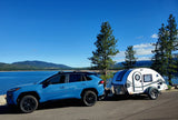 A blue SUV with an RV rental teardrop trailer parked by a beautiful lake & mountains under a clear sky.