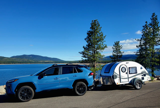 A blue SUV with an RV rental teardrop trailer parked by a beautiful lake & mountains under a clear sky.