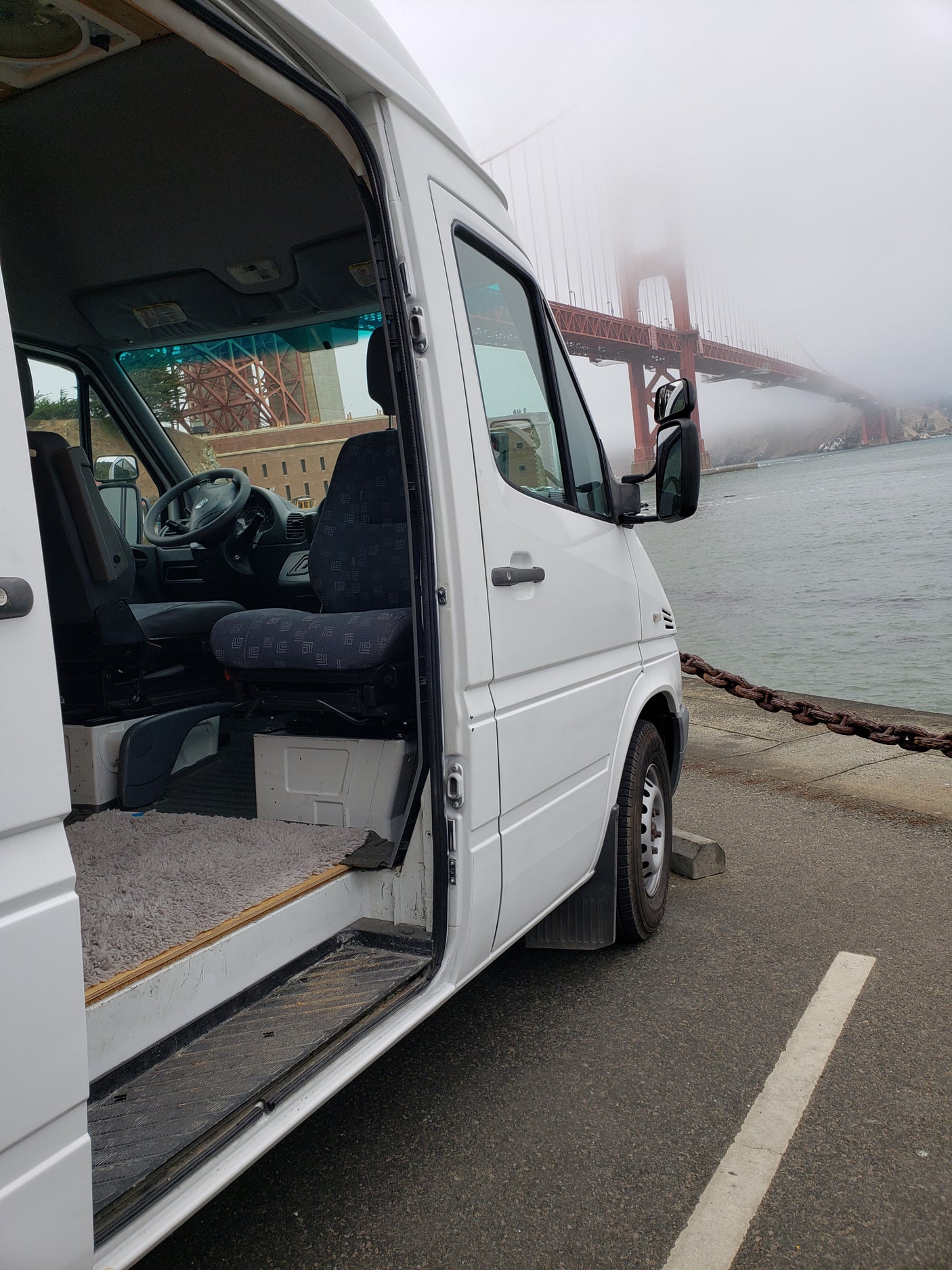 RV rental van parked with its door open, revealing the interior. The Golden Gate Bridge is visible in the fog, next to the water.