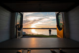RV rental view: Sunset over a tranquil lake, seen from inside a camper van with open rear doors. A person stands by the water.