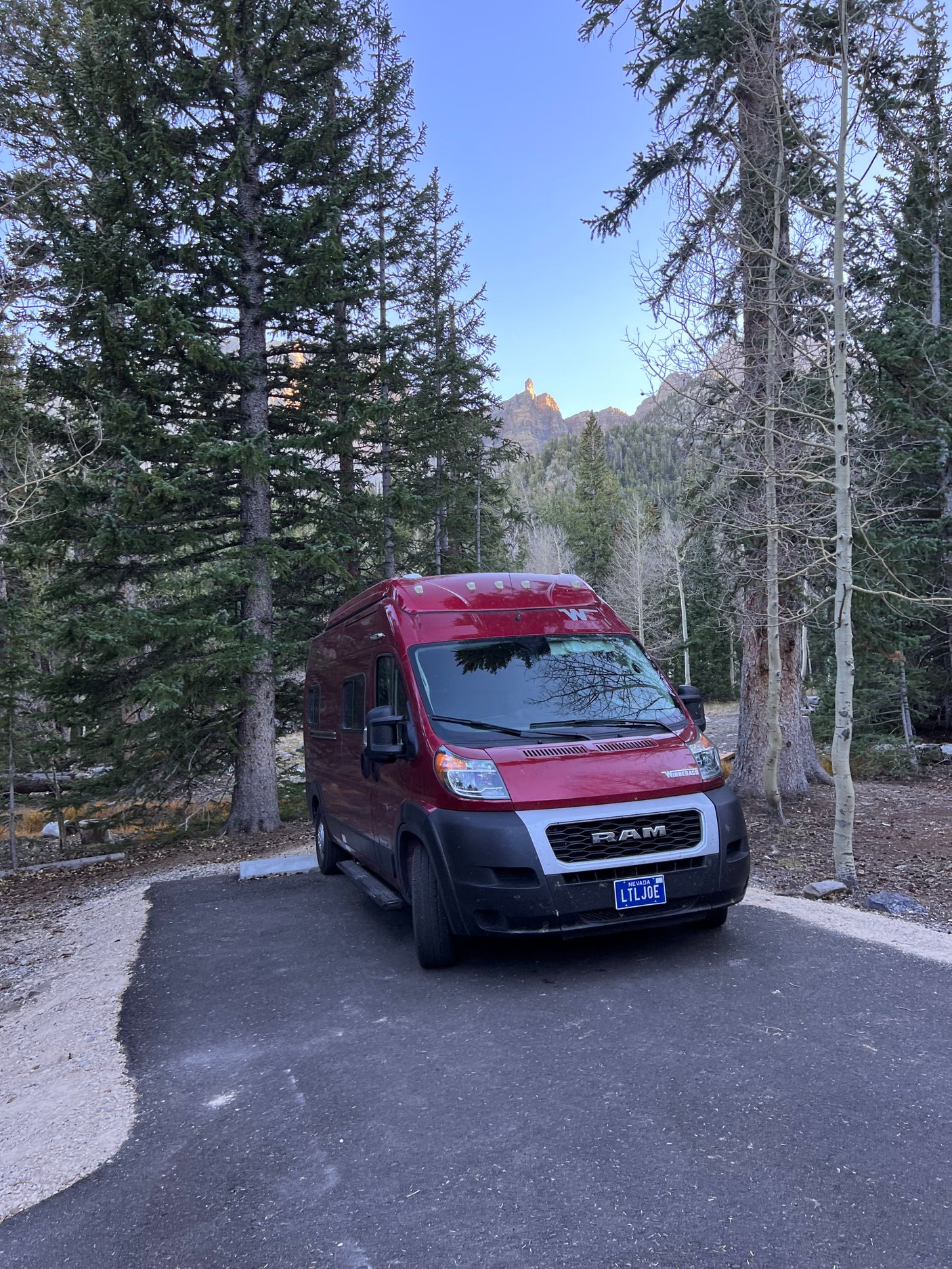 A red Winnebago camper van RV rental parked in a serene forest campsite with mountain views.