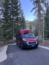 A red Winnebago camper van RV rental parked in a serene forest campsite with mountain views.