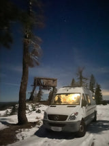 Cozy rv rental Sprinter van parked in snowy landscape at night, near a tall tree & old lookout tower.