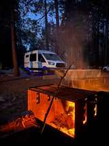 An RV rental glows blue in a dusky forest campsite, with a warm campfire in the foreground and a picnic table in the mid-ground.