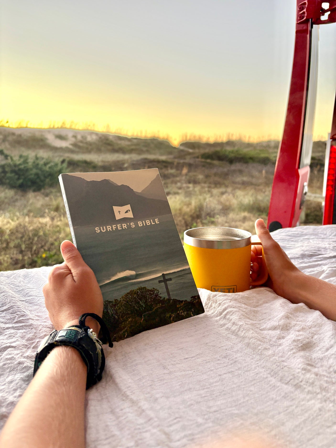 Enjoying an **rv rental** view: hands holding 'Surfer's Bible' and YETI mug, overlooking beach dunes at sunset/sunrise. A quiet spiritual retreat.