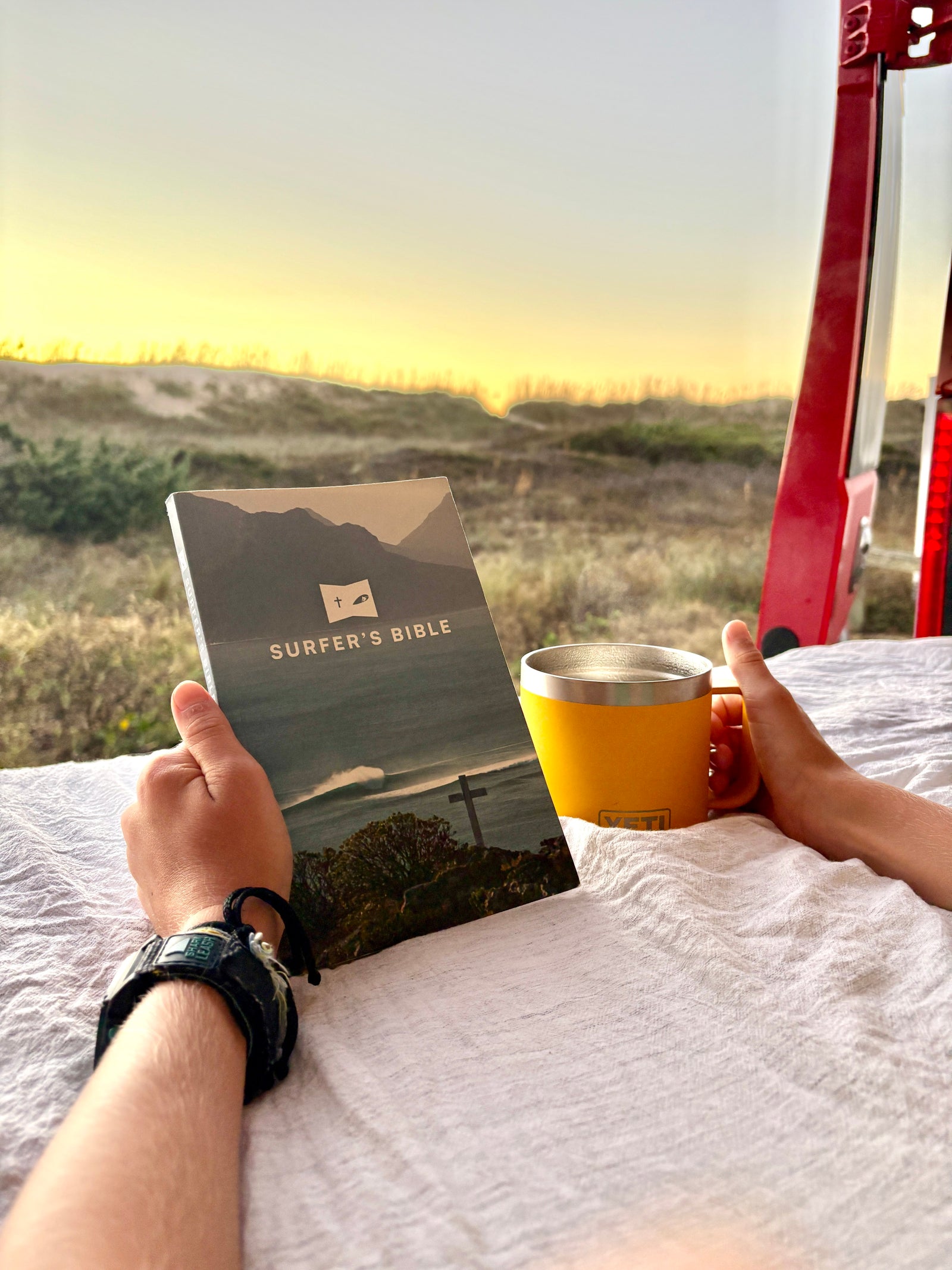 Enjoying an **rv rental** view: hands holding 'Surfer's Bible' and YETI mug, overlooking beach dunes at sunset/sunrise. A quiet spiritual retreat.