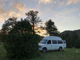 A white high-top van, resembling an RV rental, parked in a grassy field with trees at sunset, with its rear door open.