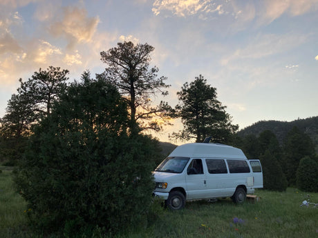 A white high-top van, resembling an RV rental, parked in a grassy field with trees at sunset, with its rear door open.