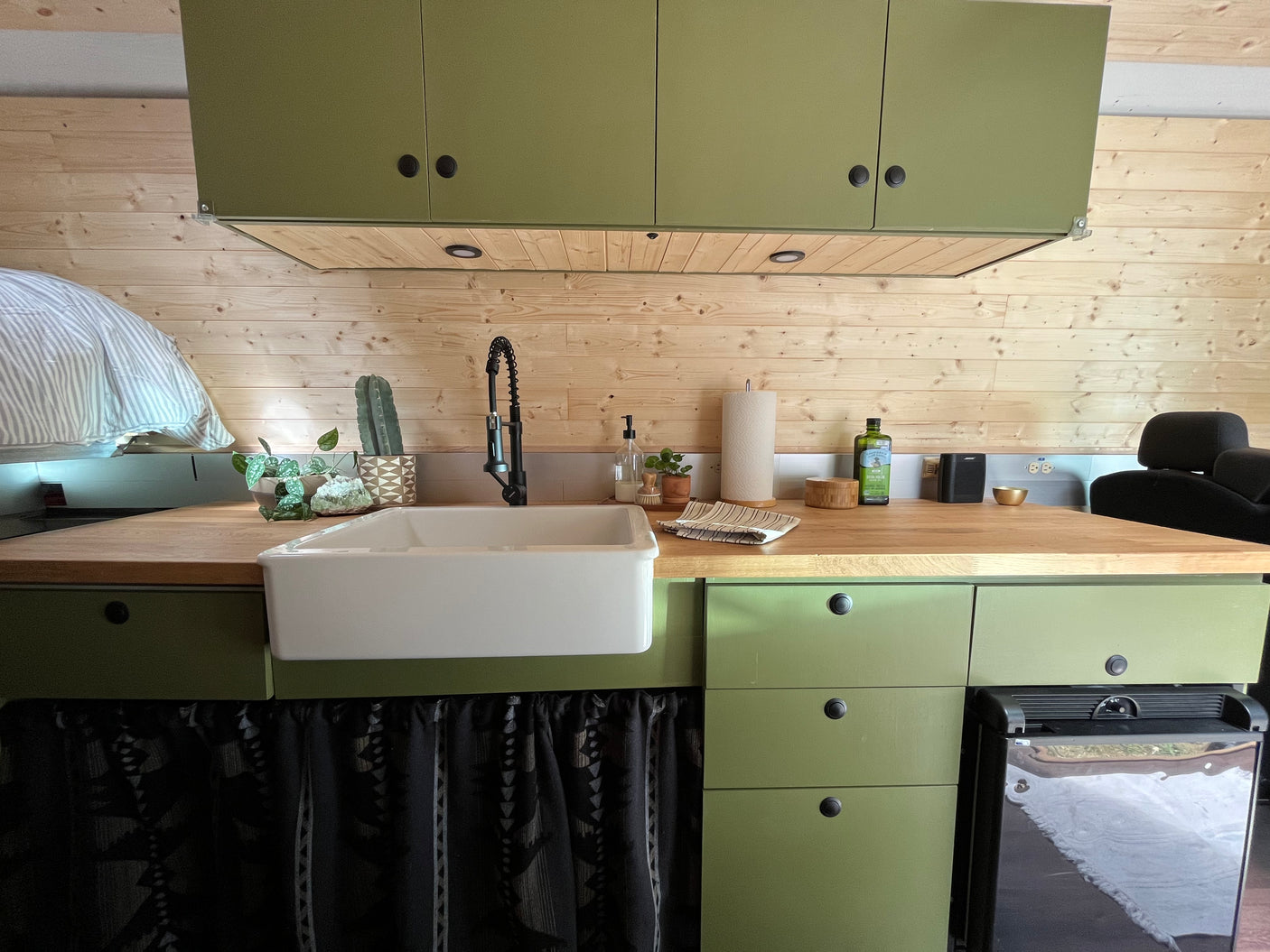 Compact RV rental kitchen with olive green cabinets, wooden walls, a white sink, and wood countertop.