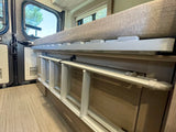 RV rental interior: A folded ladder is stored beneath a bed with a safety rail. Outside the window, trees are visible.