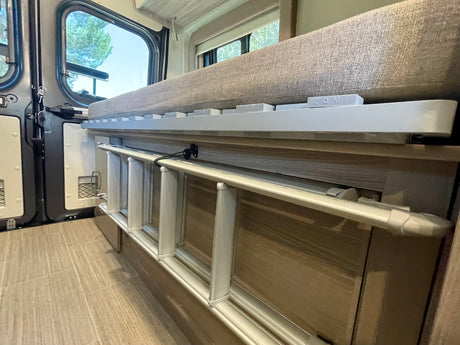 RV rental interior: A folded ladder is stored beneath a bed with a safety rail. Outside the window, trees are visible.