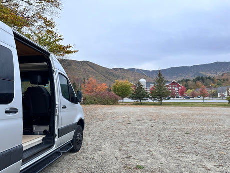 Silver RV rental campervan with open door, parked in a scenic autumn mountain setting with colorful foliage and distant ski trails.