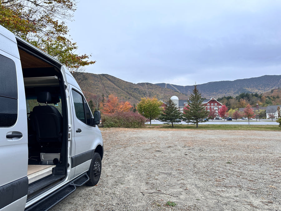 Silver RV rental campervan with open door, parked in a scenic autumn mountain setting with colorful foliage and distant ski trails.