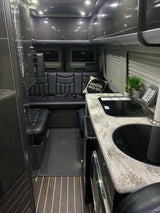 This modern RV rental interior features a functional kitchen counter, comfortable black leather seating, and overhead storage.