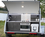 This compact RV rental features an open rear hatch revealing a fully equipped outdoor kitchen: sink, stove, microwave, and cooler.