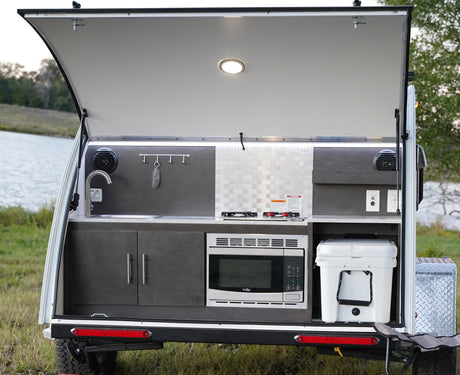 This compact RV rental features an open rear hatch revealing a fully equipped outdoor kitchen: sink, stove, microwave, and cooler.