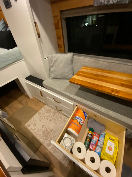RV rental interior showcases clever storage: seating, a bed, and an open drawer stocked with essentials like paper towels and toilet paper.