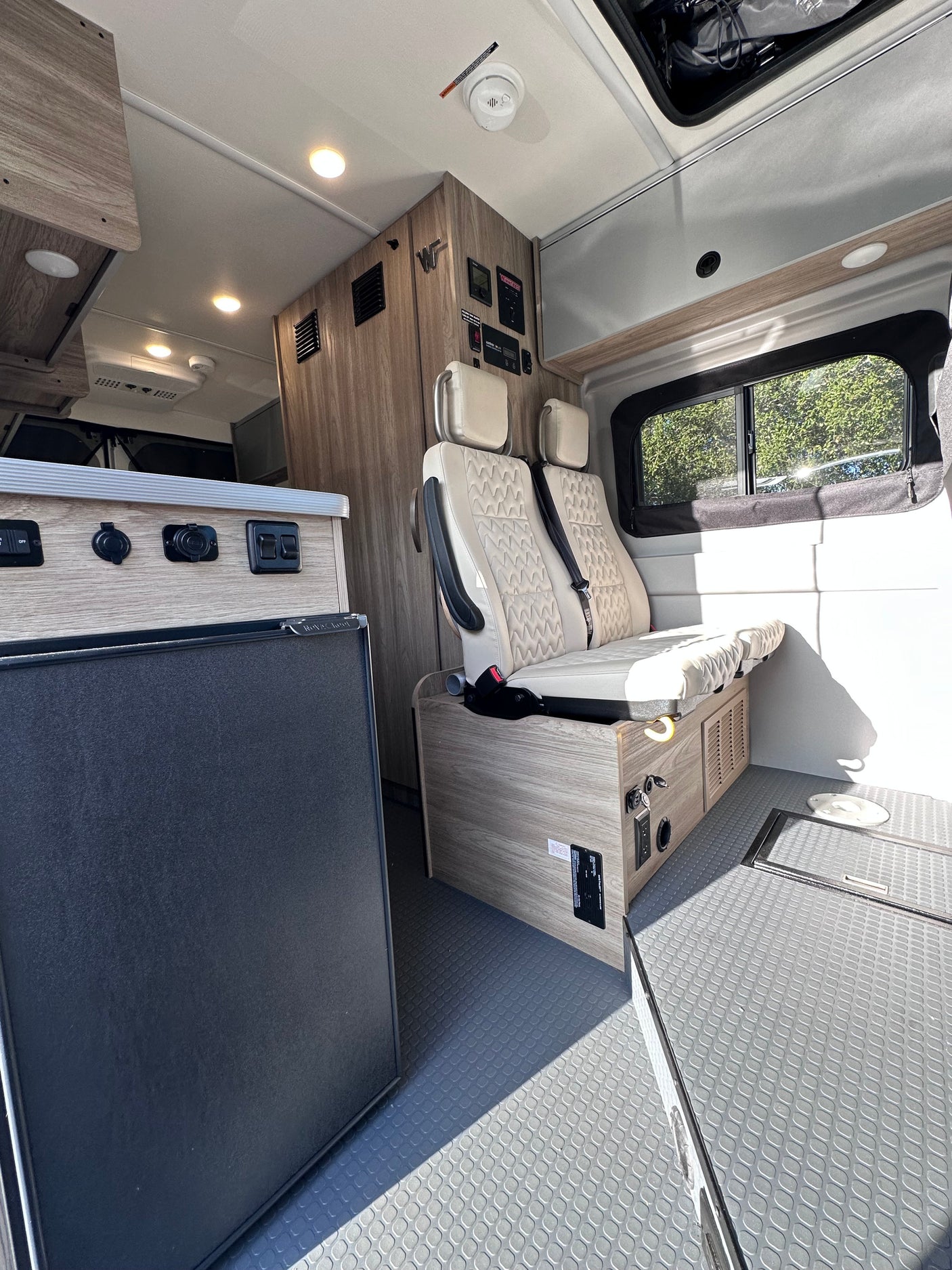 RV rental interior: Compact camper van with beige seating, a refrigerator, light wood cabinets, and control panels.