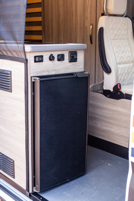 RV rental interior featuring a compact refrigerator, integrated control panel with outlets, and a comfortable passenger seat.