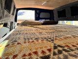 Inside an rv rental: A cozy bed with a textured blanket and pillows, looking towards the rear windows of the camper.