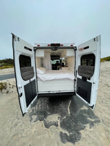 Rear doors of a white camper van open on sand, revealing a cozy bed inside. Ideal RV rental for your next beach adventure.