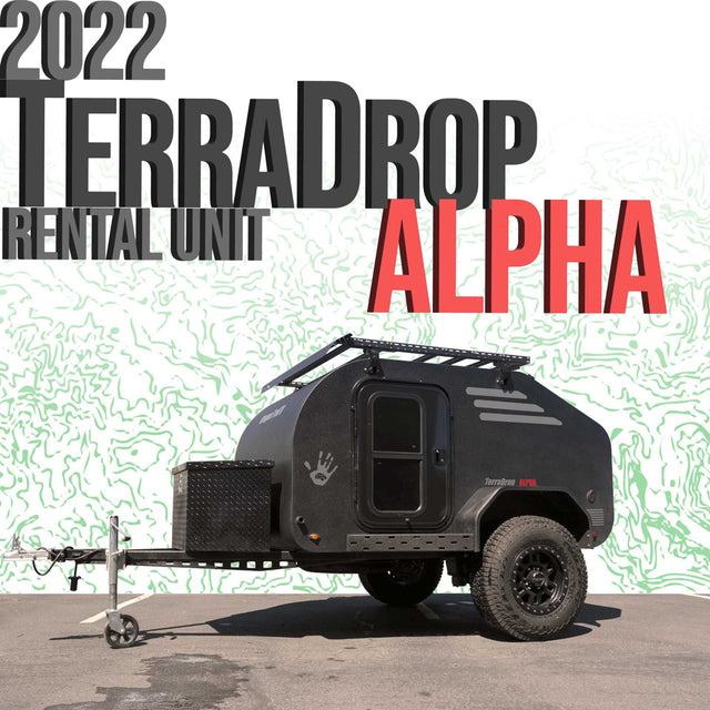 This 2022 TerraDrop Alpha teardrop RV rental unit is a black trailer with off-road tires and a roof rack, parked on asphalt.
