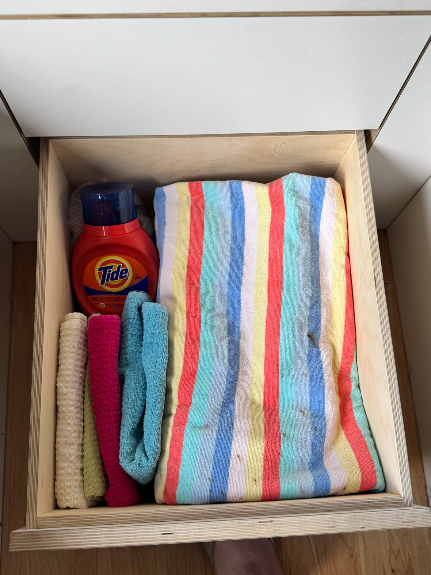 Compact RV rental storage: an open drawer holds Tide detergent, various cleaning cloths, and a striped towel.